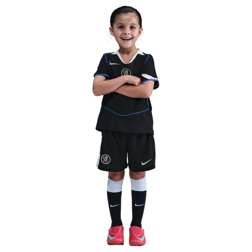Young girl smiling with arms crossed, wearing a black Chelsea FC soccer kit and pink cleats against a solid black background.