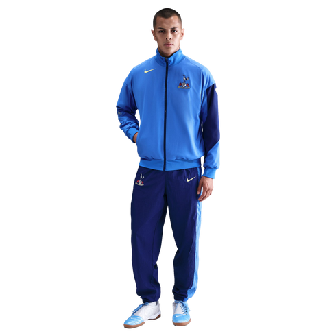 A man wearing a light blue and navy Nike Tottenham Hotspur FC tracksuit and matching sneakers, standing against a solid black background.