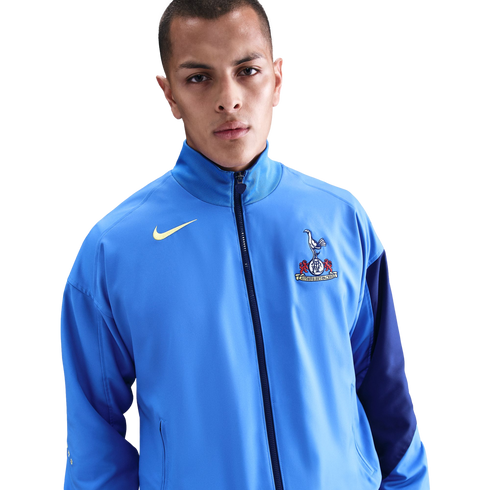 A man wearing a light blue Nike Tottenham Hotspur track jacket against a solid black background.