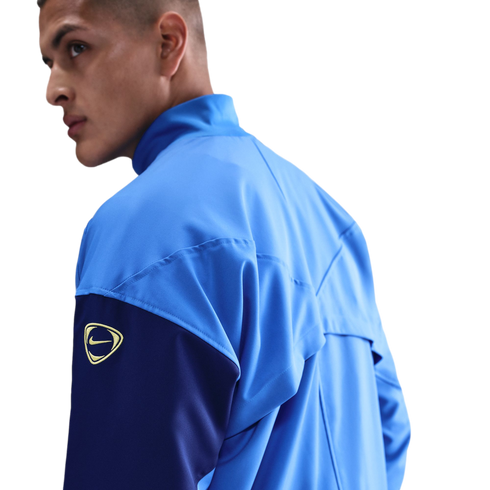 A man looking over his shoulder in a light blue and navy Nike jacket with a gold logo on the arm, set against a black background.
