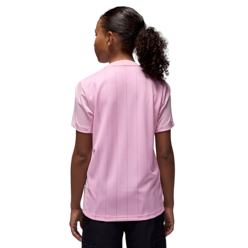 Rear view of a person with a curly ponytail wearing a light pink, pinstriped t-shirt and black shorts against a black background.