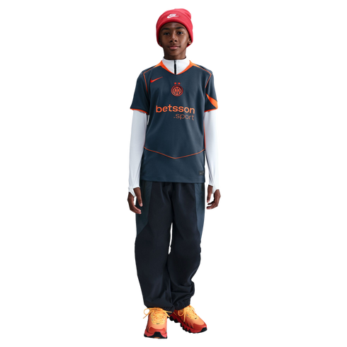 Young person in a red Nike beanie, grey and orange Inter Milan jersey, dark pants, and orange sneakers standing against a black background.