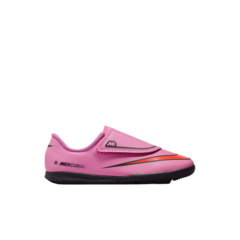 A bright pink Nike Mercurial indoor soccer shoe for kids, featuring a black outsole, an orange Nike swoosh, and a hook-and-loop strap closure.