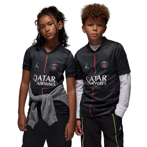 A young girl and boy wear matching black Paris Saint-Germain soccer jerseys with red vertical stripes and logos while posing against a solid black background.