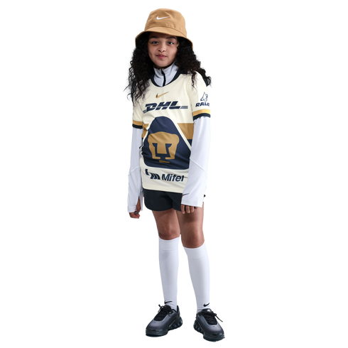 A young girl with long curly hair wearing a soccer jersey, tan bucket hat, black shorts, and white knee-high socks, standing against a plain white background.
