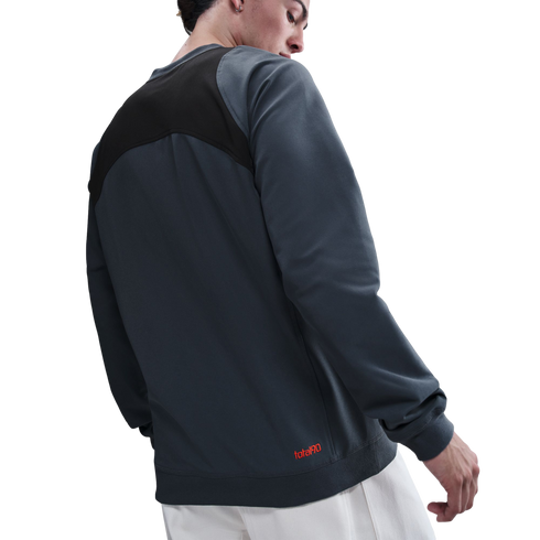 A man seen from behind wearing a grey and black color-blocked sweatshirt with a red logo on the hem and white pants.