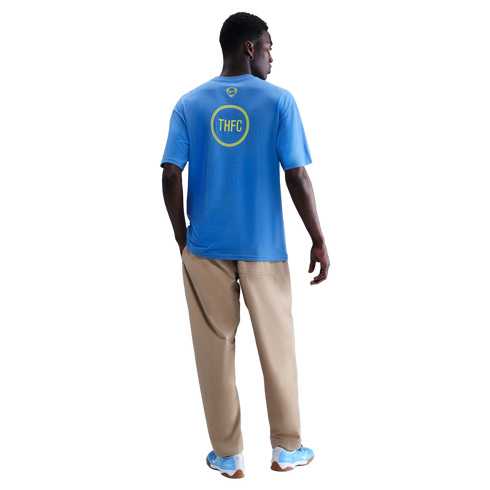 Back view of a man wearing a blue Nike t-shirt with a yellow "THFC" graphic, tan pants, and blue sneakers against a black background.