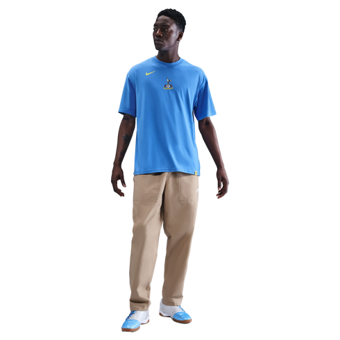 Full-length portrait of a man wearing a light blue Nike Tottenham Hotspur t-shirt, tan trousers, and blue and white sneakers, standing against a solid black background.