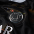 Close-up of a black sports jersey featuring a metallic silver Paris Saint-Germain club crest. The fabric has an abstract, multicolored pattern with hints of red and green.