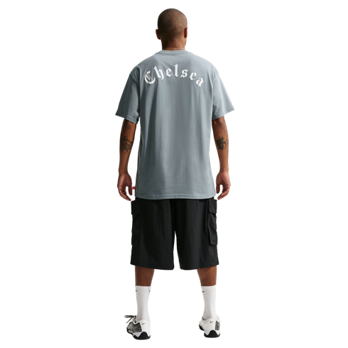 Back view of a man wearing a grey t-shirt with "Chelsea" in white gothic font, black cargo shorts, white socks, and sneakers against a black background.