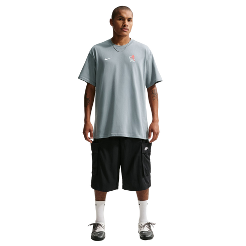 Full-length shot of a man in a grey Nike t-shirt, black cargo shorts, white socks, and grey shoes against a black background. He has a necklace and arm tattoos.