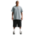 Full-length shot of a man in a grey Nike t-shirt, black cargo shorts, white socks, and grey shoes against a black background. He has a necklace and arm tattoos.