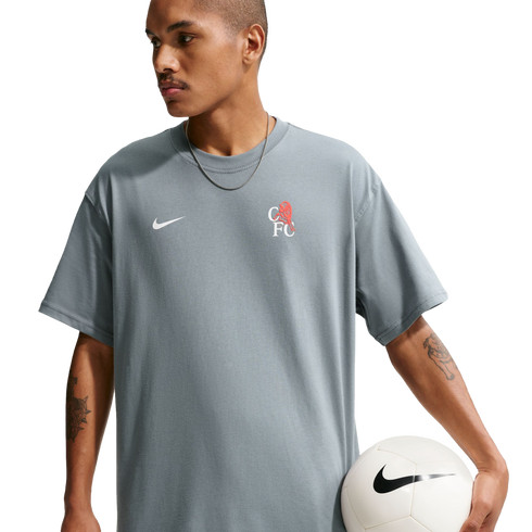 A person with arm tattoos wearing a grey Nike Chelsea FC t-shirt holds a soccer ball against a black background.