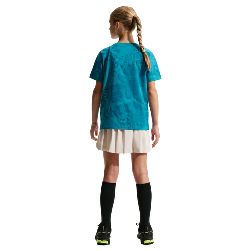 Back view of a girl with a blonde braid wearing a teal patterned t-shirt, beige pleated skirt, black knee-high socks, and black sneakers against a black background.