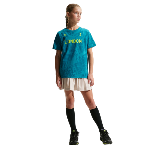 A young girl in a teal "LONDON" t-shirt, beige pleated skirt, and black sneakers poses against a black background.