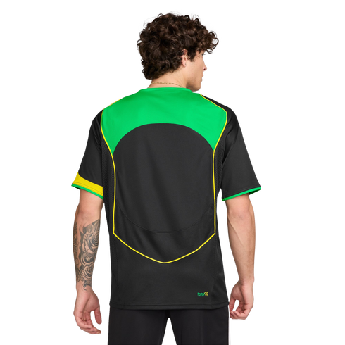 Rear view of a man with curly hair wearing a black soccer jersey with green and yellow accents. He has a tattoo on his left forearm. Plain white background.