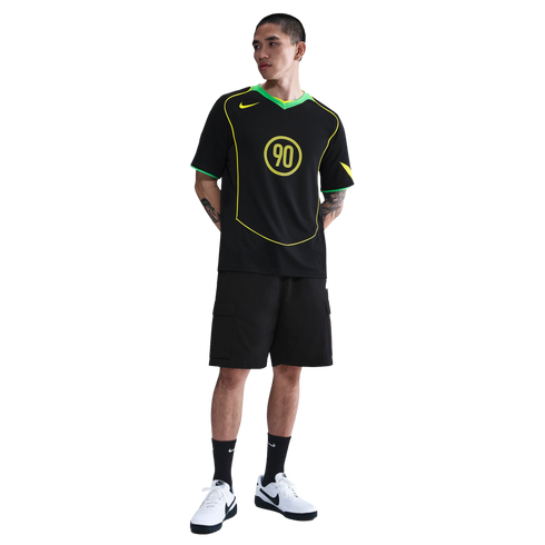 A man poses in a black Nike T90 soccer jersey, black cargo shorts, and white sneakers against a white background.