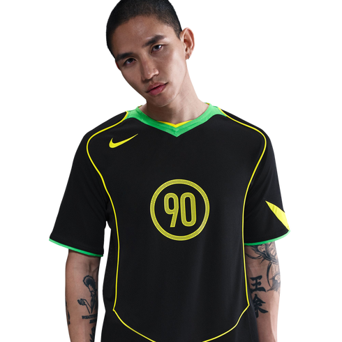 A man with arm tattoos wears a black Nike Total 90 jersey with neon green and yellow accents, set against a plain white background.