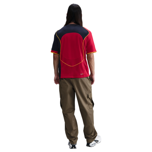 Back view of a person with long dark hair wearing a red, black, and yellow jersey, olive green cargo pants, and white and black sneakers on a white background.