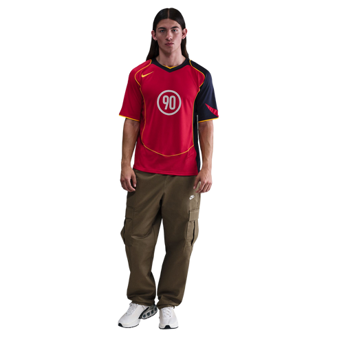 Man with long dark hair wearing a red Nike Total 90 jersey, olive green cargo pants, and white sneakers.