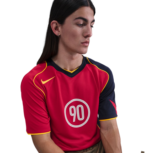 A person with long dark hair wearing a red and navy Nike Total 90 sports jersey with yellow trim, looking down against a white background.