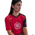 A person with long dark hair wearing a red and navy Nike Total 90 sports jersey with yellow trim, looking down against a white background.