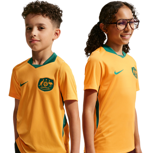A boy and a girl pose side-by-side against a black background wearing gold Nike Australia national soccer team jerseys with green trim. The boy looks towards the camera while the girl, wearing glasses, smiles and looks off to the right.