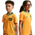 A boy and a girl pose side-by-side against a black background wearing gold Nike Australia national soccer team jerseys with green trim. The boy looks towards the camera while the girl, wearing glasses, smiles and looks off to the right.