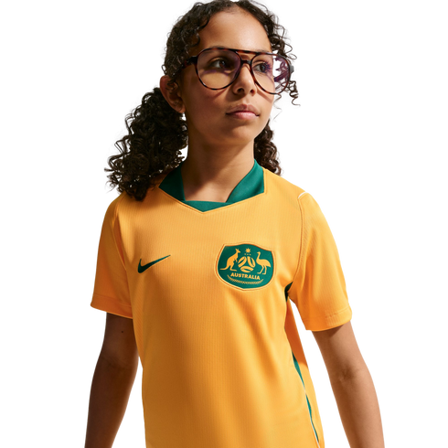 A waist-up portrait of a young girl with curly dark hair and glasses, wearing a yellow and green Australian national soccer team jersey against a black background.