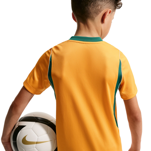 A rear view of a child wearing a gold soccer jersey with green trim, holding a white soccer ball under their arm against a solid black background.