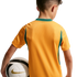A rear view of a child wearing a gold soccer jersey with green trim, holding a white soccer ball under their arm against a solid black background.