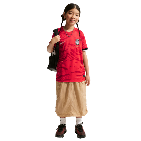 A smiling girl with braids wears a red Korea soccer jersey, tan skirt, and black backpack against a black background.