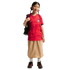 A smiling girl with braids wears a red Korea soccer jersey, tan skirt, and black backpack against a black background.