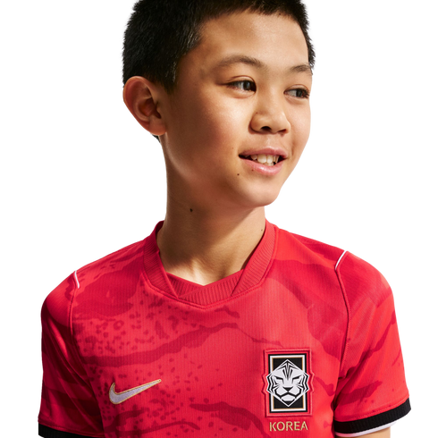 A smiling young boy wearing a red South Korea national football jersey with a subtle tiger pattern, against a solid black background.