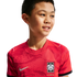 A smiling young boy wearing a red South Korea national football jersey with a subtle tiger pattern, against a solid black background.