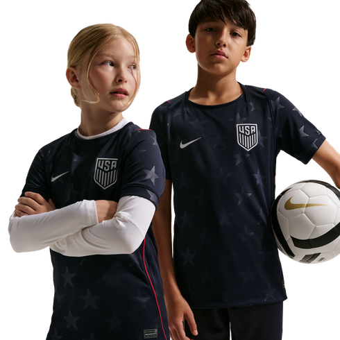 A girl and boy wear navy blue star-patterned USA soccer jerseys; the boy holds a soccer ball. Black background.