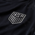 A close-up of a dark ribbed jersey featuring an embroidered silver U.S. Soccer Federation shield crest with "USA" and vertical stripes.