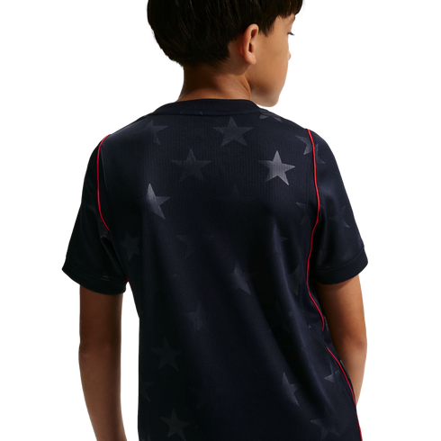 Rear view of a child wearing a navy blue short-sleeved t-shirt with a tonal star pattern and red piping along the shoulders.