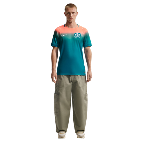 A man standing against a black background wearing a Nike Australia national team jersey with a peach-to-teal gradient and oversized olive green cargo pants.