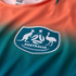 A close-up of a sports jersey featuring an orange-to-teal gradient fabric with vertical ribbing. Centered is a teal shield-shaped crest with white embroidery of a kangaroo, an emu, a stylized soccer ball, and the word "AUSTRALIA."