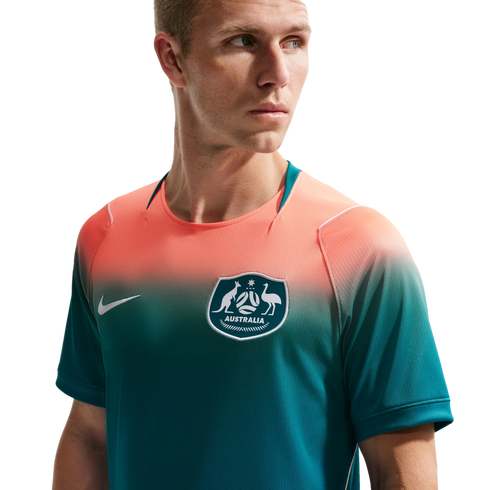 A man wearing an Australian national soccer team jersey with a salmon-to-teal color gradient, featuring the team crest and a white Nike logo. He is looking off-camera against a solid black background.