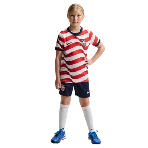 A young girl in a red and white wavy striped USA soccer uniform, navy shorts, and bright blue cleats, standing against a solid black background.