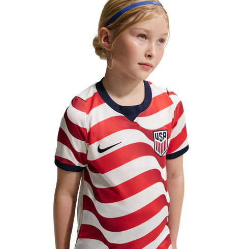 A young girl in a red and white striped USA soccer jersey and blue headband, set against a black background.