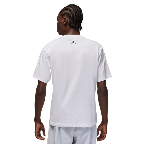 A rear view of a man with braided hair wearing a white t-shirt with a small black logo on the back, set against a black background.