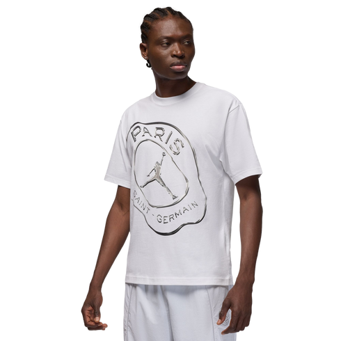 A person with braided hair wears a white T-shirt featuring a silver Jordan x PSG graphic, paired with white shorts against a solid black background.