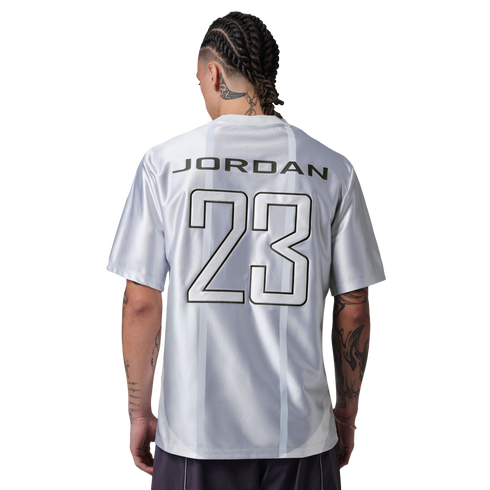 A person with braided hair and tattoos stands with their back to the camera, wearing a silver jersey with "JORDAN 23" printed on the back against a black background.