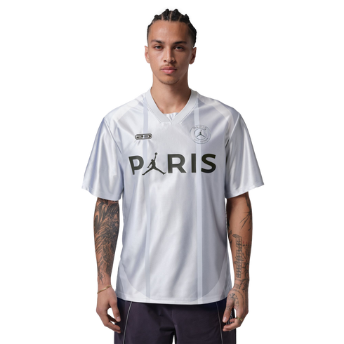 A man with arm tattoos wearing a white Jordan Paris Saint-Germain jersey with "PARIS" across the chest, standing against a black background.