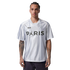 A man with arm tattoos wearing a white Jordan Paris Saint-Germain jersey with "PARIS" across the chest, standing against a black background.