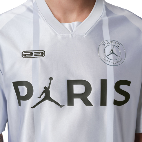 Close-up of a white PSG jersey featuring a large black 'PARIS' graphic with a Jumpman logo replacing the letter 'A', faint grey stripes, and team crest.