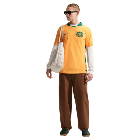 A man in a yellow Australia soccer jersey, white long-sleeved shirt, brown pants, and green sneakers carries a white tote bag against a black background.